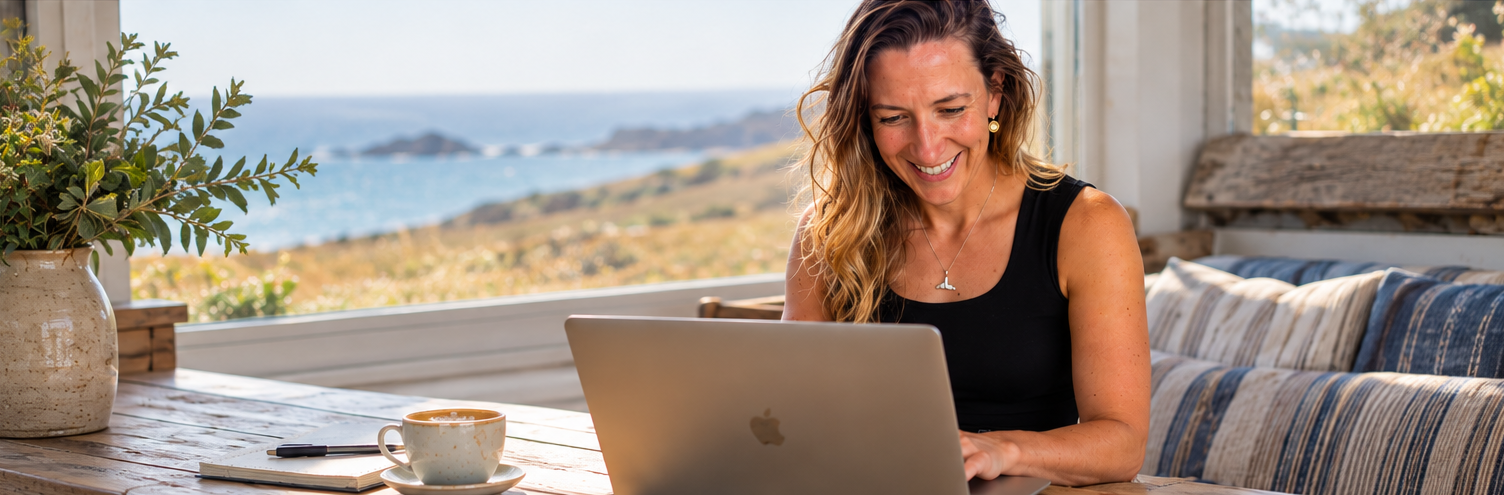 Bronwyn Lear, founder of WildKind Collective, smiling at her laptop with coffee, the coastline behind her